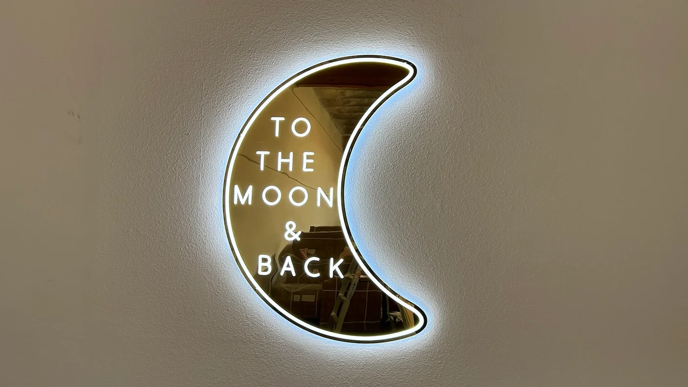 Neon sign in the shape of a crescent moon with the phrase "TO THE MOON & BACK" written inside. The sign emits a soft white glow, mounted on a plain wall, creating a romantic and whimsical decorative piece.