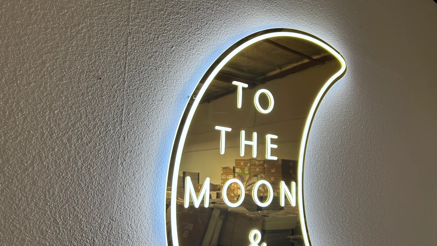 Neon sign in the shape of a crescent moon with the phrase "TO THE MOON & BACK" written inside. The sign emits a soft white glow, mounted on a plain wall, creating a romantic and whimsical decorative piece.