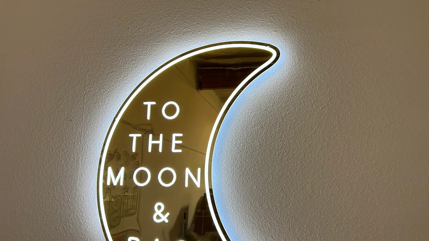 Neon sign in the shape of a crescent moon with the phrase "TO THE MOON & BACK" written inside. The sign emits a soft white glow, mounted on a plain wall, creating a romantic and whimsical decorative piece.