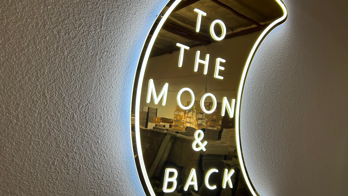 Neon sign in the shape of a crescent moon with the phrase "TO THE MOON & BACK" written inside. The sign emits a soft white glow, mounted on a plain wall, creating a romantic and whimsical decorative piece.