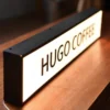 Illuminated "COFFEE" light box sign placed on a wooden surface, used as business signage for a coffee shop. The sign has a sleek black frame and emits a warm glow, creating a cozy ambiance.
