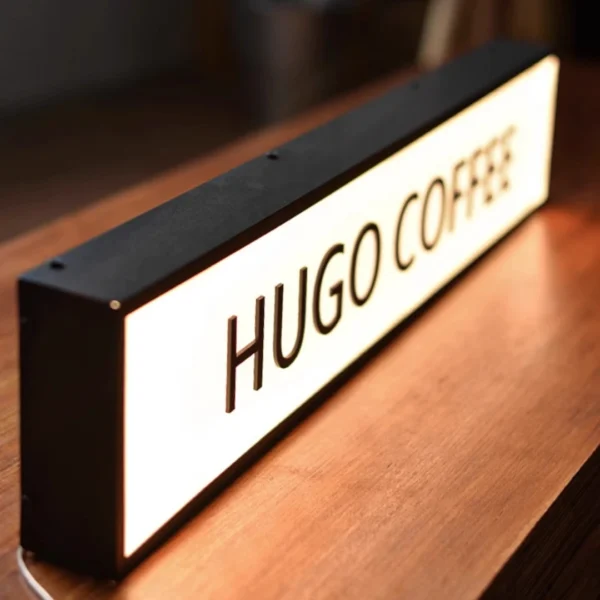 Illuminated "COFFEE" light box sign placed on a wooden surface, used as business signage for a coffee shop. The sign has a sleek black frame and emits a warm glow, creating a cozy ambiance.