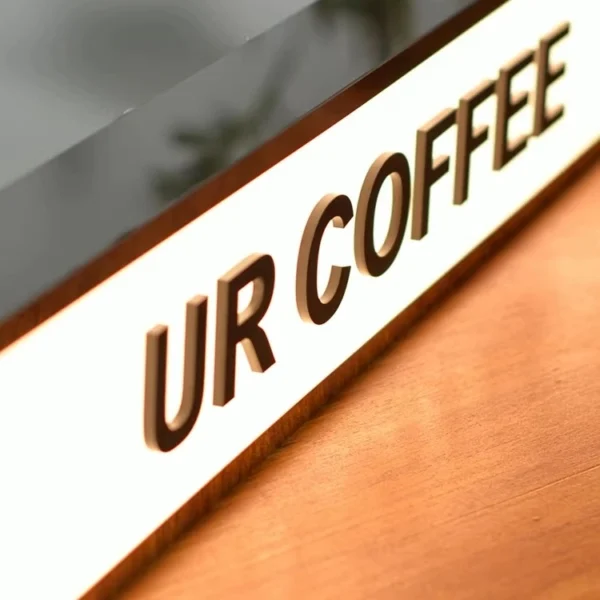 Illuminated "COFFEE" light box sign placed on a wooden surface, used as business signage for a coffee shop. The sign has a sleek black frame and emits a warm glow, creating a cozy ambiance.