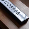 Illuminated "COFFEE" light box sign placed on a wooden surface, used as business signage for a coffee shop. The sign has a sleek black frame and emits a warm glow, creating a cozy ambiance.