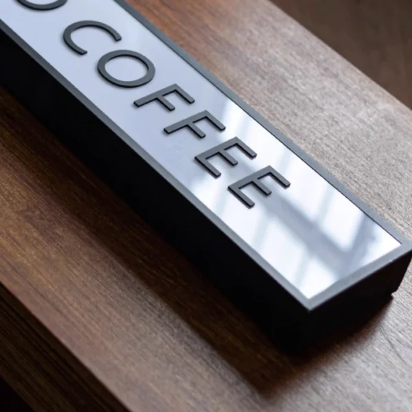 Illuminated "COFFEE" light box sign placed on a wooden surface, used as business signage for a coffee shop. The sign has a sleek black frame and emits a warm glow, creating a cozy ambiance.