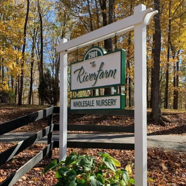 Outdoor signage mounted on a white wooden frame. The sign features a green and white color scheme with a cursive font for the nursery's name and block letters for the rest.