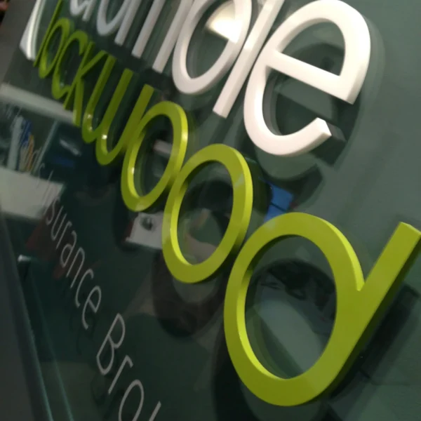 A close-up view of a wall sign featuring the words in white and green 3D letters. The sign is mounted on a glass or glossy dark surface, with a slight reflection visible. The modern design and color contrast make the sign visually striking.
