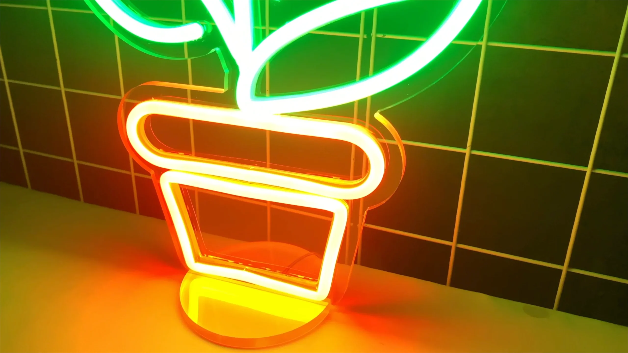 A desktop neon sign depicts a green-leaf plant and an orange flowerpot. The sign is placed on a desk, emitting vibrant light, and can be dimmed. It can also serve as a night light and an ambient light.